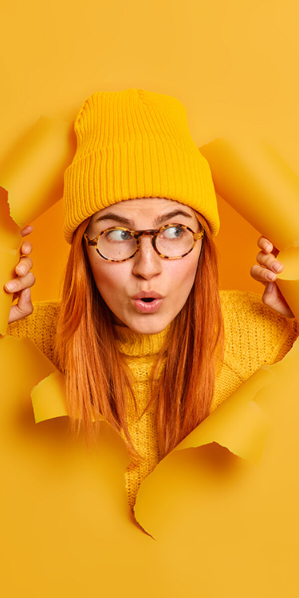 Beautiful surprised redhead beautiful woman has wondered face expression opens mouth stares at something unbelievable looks through hole in yellow paper background wears hat transparent glasses