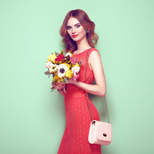 Blonde young woman in elegant red dress. Girl posing on a green background with handbag. Jewelry and hairstyle. Lady with spring bouquet of flowers. Fashion photo