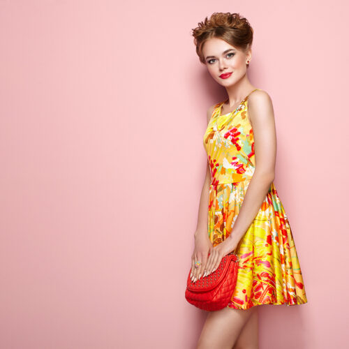Portrait of Fashion Young woman in Floral Dress. Female Model in Stylish Spring Summer Outfit. Girl Posing on a Pink Background. Stylish Hairstyle. Blonde Lady with Red Handbag