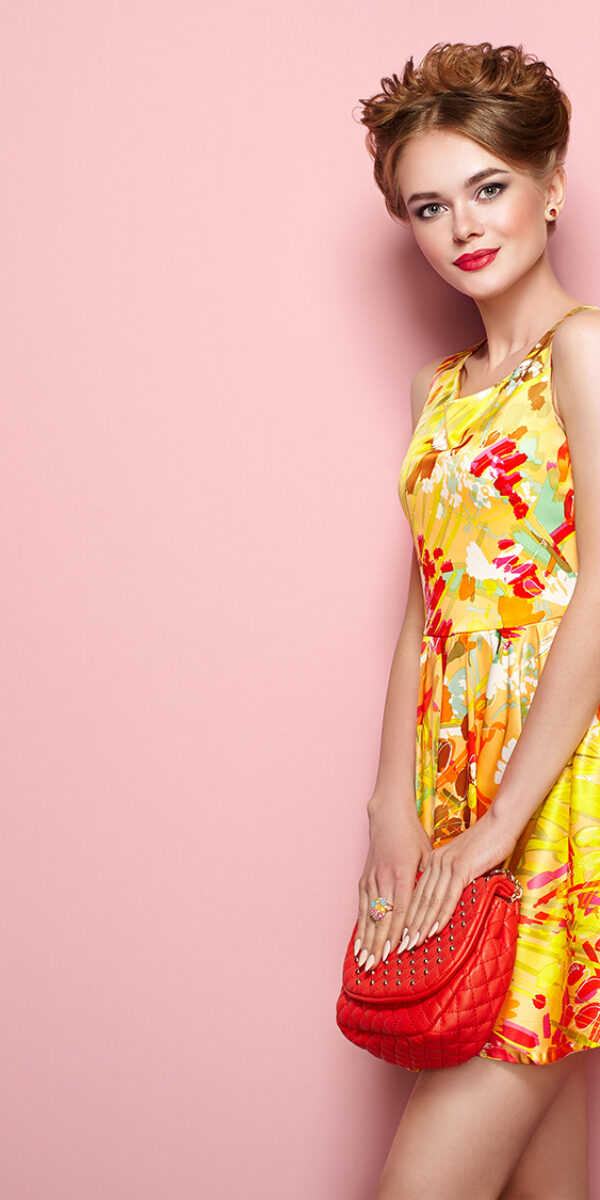 Portrait of Fashion Young woman in Floral Dress. Female Model in Stylish Spring Summer Outfit. Girl Posing on a Pink Background. Stylish Hairstyle. Blonde Lady with Red Handbag