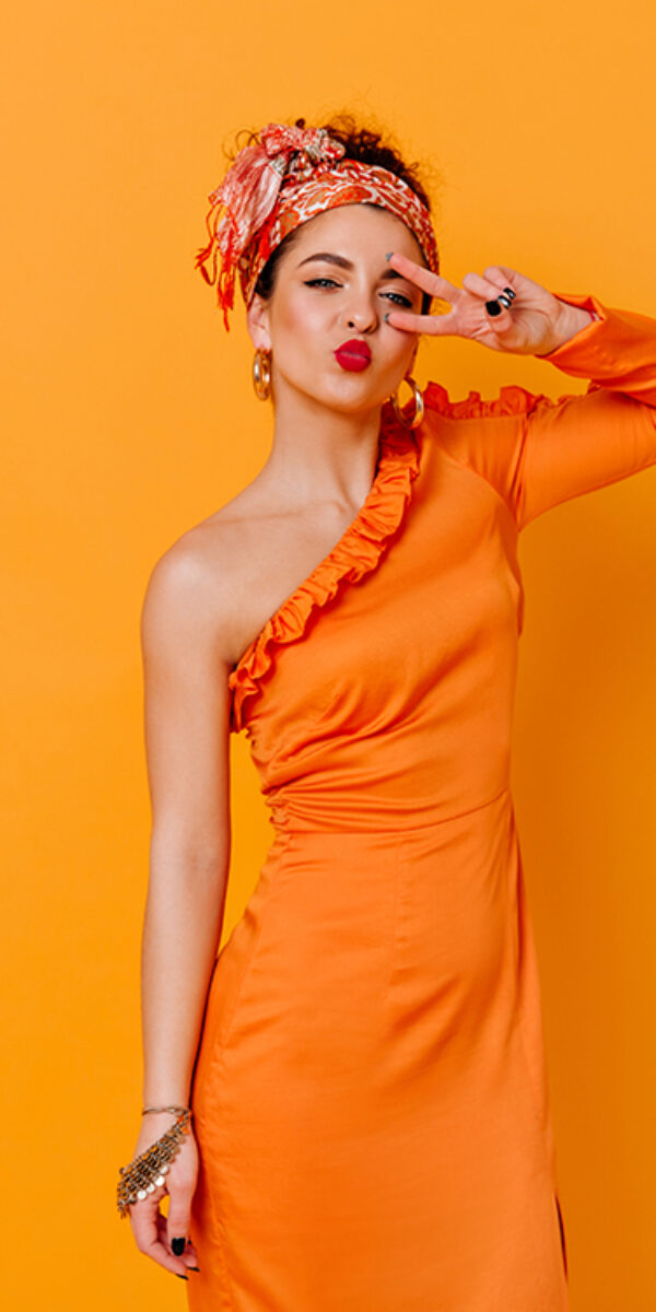 Coquettish woman in silk long dress and headband blows kiss and shows peace sign on orange background