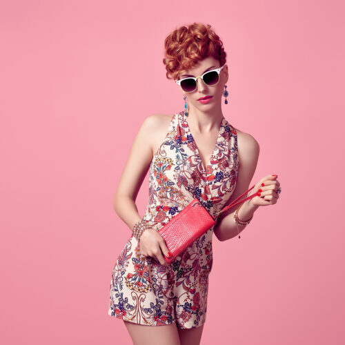 Fashion Model in Sexy Jumpsuit. Stylish Mohawk hairstyle, fashion Sunglasses, Summer Floral Outfit. Beauty Redhead Woman in Trendy Summer Dress. Glamour fashion Clutch. Playful Luxury Girl on Pink