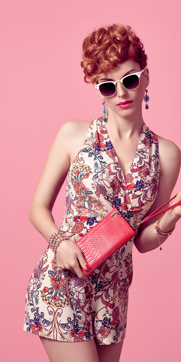 Fashion Model in Sexy Jumpsuit. Stylish Mohawk hairstyle, fashion Sunglasses, Summer Floral Outfit. Beauty Redhead Woman in Trendy Summer Dress. Glamour fashion Clutch. Playful Luxury Girl on Pink