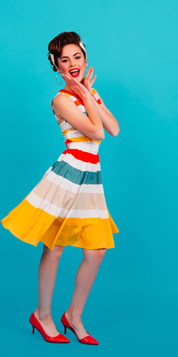 Full length view of pinup girl in striped dress. Studio shot of brunette lady dancing on blue backg Full length view of pinup girl in striped dress. Studio shot of brunette lady dancing on blue background.