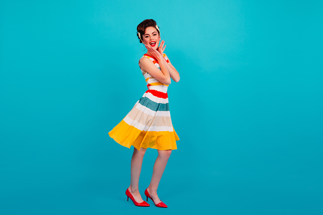 Full length view of pinup girl in striped dress. Studio shot of brunette lady dancing on blue backg