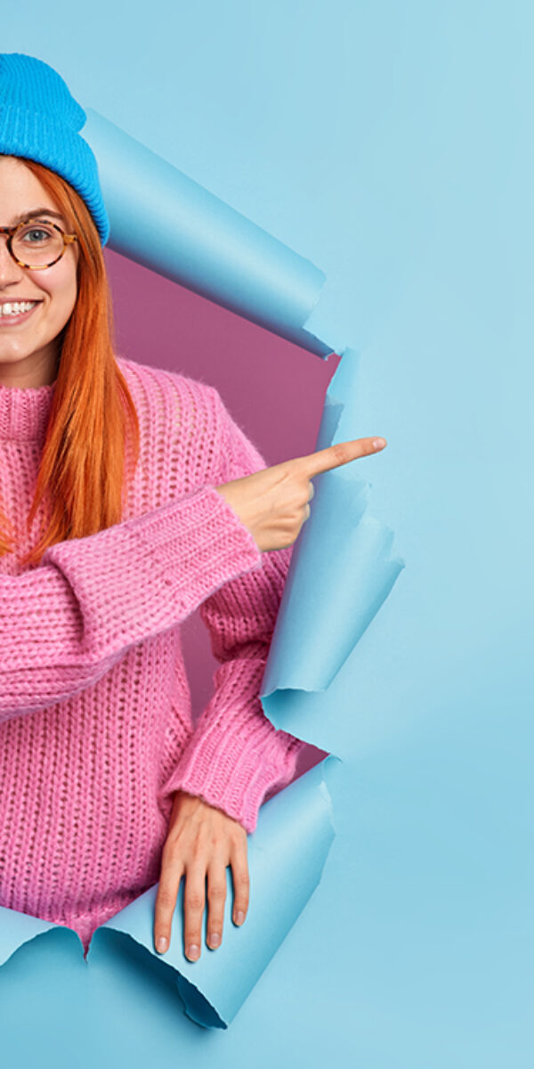 Happy beautiful redhead young woman points right at copy space blank for promo banner wears blue hat and knitted blue jumper suggests follow page to find out information poses in ripped hole Happy beautiful redhead young woman points right at copy space blank for promo banner wears blue hat and knitted blue jumper suggests follow page to find out information poses in ripped hole