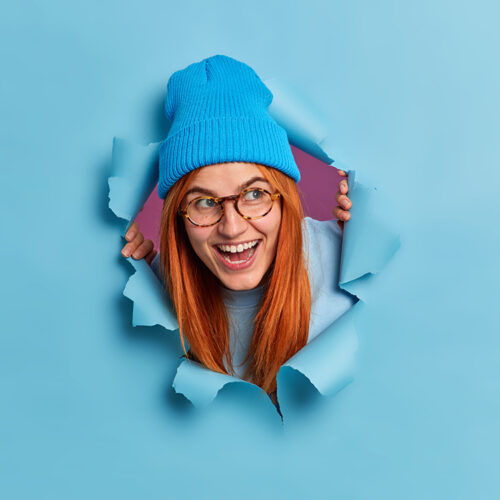 Happy redhead young woman giggles positively and looks aside has fun feels entertained wears optical glasses blue hat looks through torn paper background. Joyful foxy female teenager indoor. Happy redhead young woman giggles positively and looks aside has fun feels entertained wears optical glasses blue hat looks through torn paper background. Joyful foxy female teenager indoor.