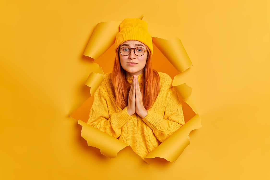 Hopleless sad woman has imploring expression keeps palms together asks for apology wears yellow hat and sweater poses through paper hole of yellow background. Please help me or give chance to improve