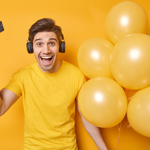 Horizontal shot of cheerful young man holds plastic card and bunch of inflated balloons smiles gladfully celebrates birthday spends money on preparing for party wears casual t shirt listens music