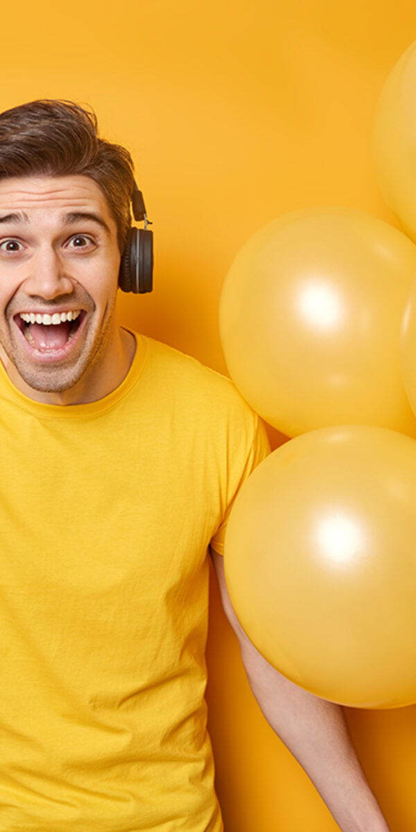 Horizontal shot of cheerful young man holds plastic card and bunch of inflated balloons smiles gladfully celebrates birthday spends money on preparing for party wears casual t shirt listens music