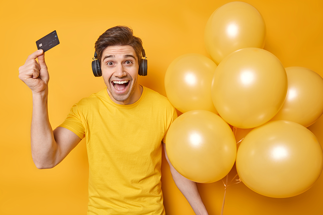 Horizontal shot of cheerful young man holds plastic card and bunch of inflated balloons smiles gladfully celebrates birthday spends money on preparing for party wears casual t shirt listens music