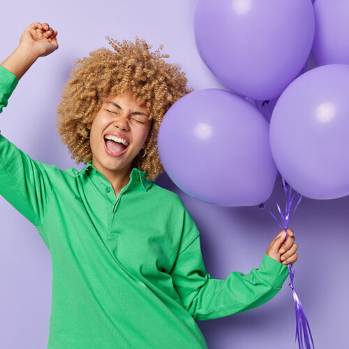 Horizontal shot of overjoyed woman keeps palm raised up holds bunch on inflated balloons dressed in green jumper isolated over purple background celebrates anniversary. Happy birthday concept