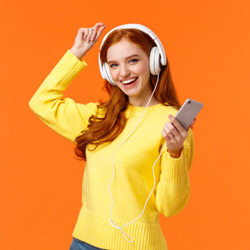 Lets dance. Cheerful carefree modern hipster girl with red curly hair and freckles, dancing lift hand up and holding smartphone, listen music with new ordered online headphones, orange background.