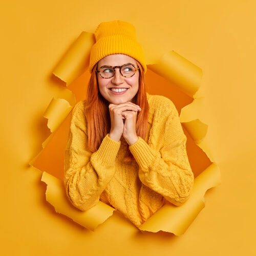 Lovely young female model with red hair keeps hands under chin concentrated aside and smiles gently wears fashionable youth clothes thinks about future plans imagines something good in mind.