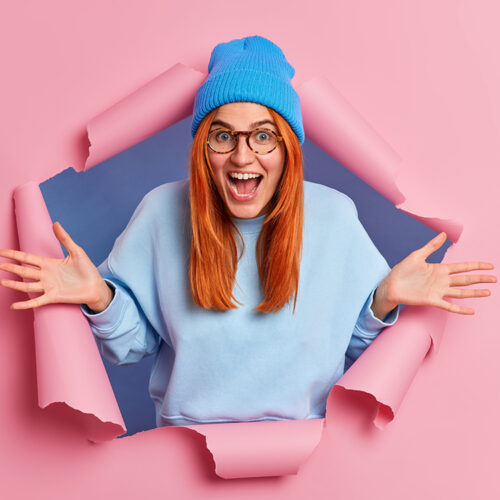Overemotive positive ginger young woman exclaims loudly spreads palms feels very glad wears blue hat and jumper breaks through pink paper background opens mouth from amazement. Wow how nice.