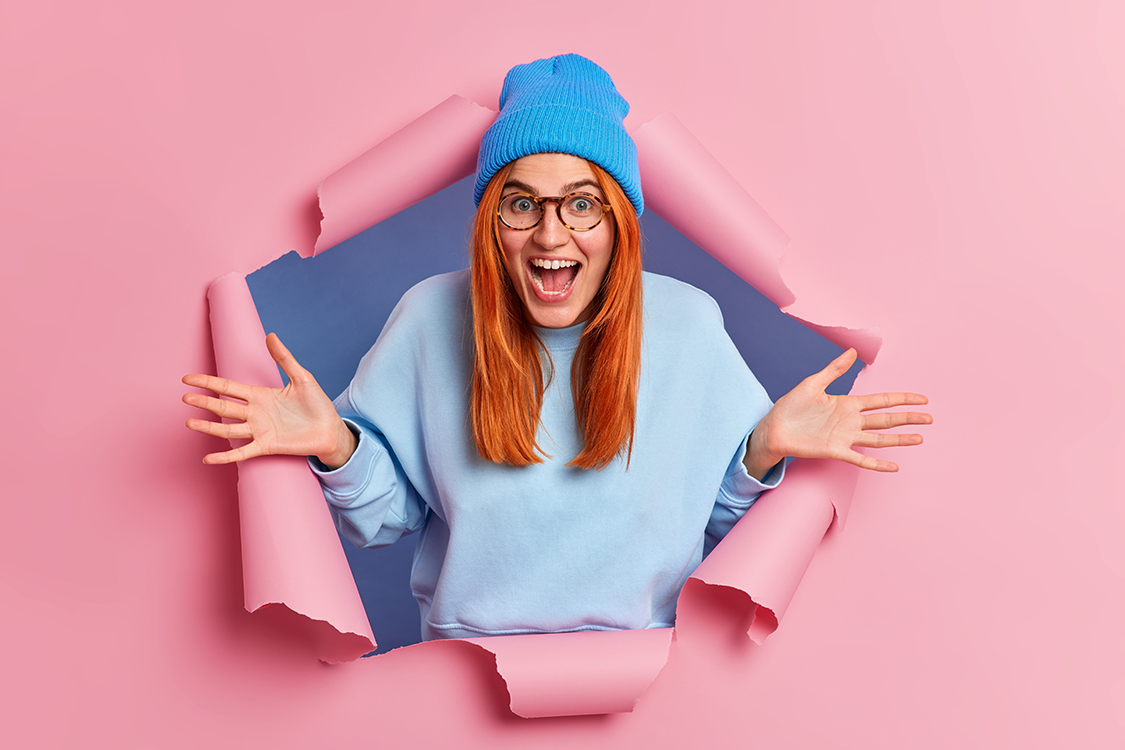 Overemotive positive ginger young woman exclaims loudly spreads palms feels very glad wears blue hat and jumper breaks through pink paper background opens mouth from amazement. Wow how nice.