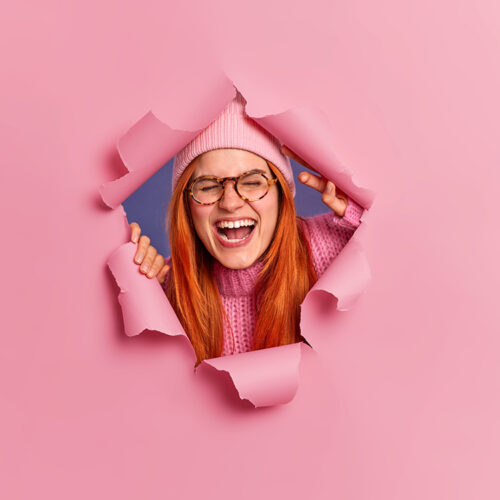 Overjoyed redhead freckled woman wears glasses and hat poses through ripped hole laughs joyfully keeps mouth opened keeps eyes closed. Satisfied foxy European girl hears something very funny Overjoyed redhead freckled woman wears glasses and hat poses through ripped hole laughs joyfully keeps mouth opened keeps eyes closed. Satisfied foxy European girl hears something very funny
