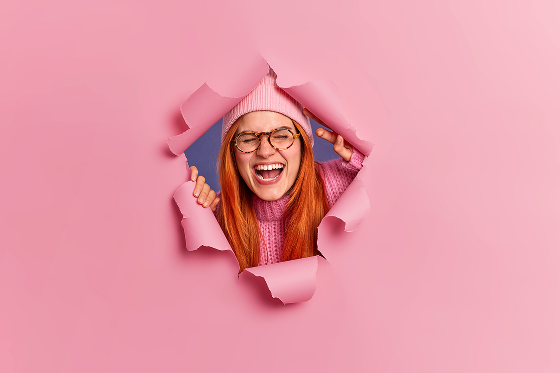 Overjoyed redhead freckled woman wears glasses and hat poses through ripped hole laughs joyfully keeps mouth opened keeps eyes closed. Satisfied foxy European girl hears something very funny
