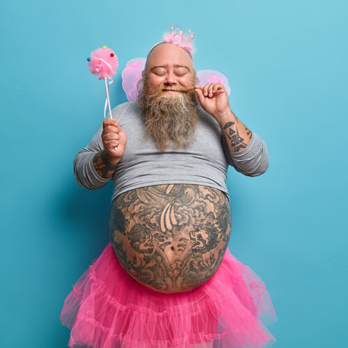 Positive funny bearded male princess holds magic wand and entertains guests on costume party shows fat abdomen poses against blue background. Cheerful daddy has image of fairy plays with children