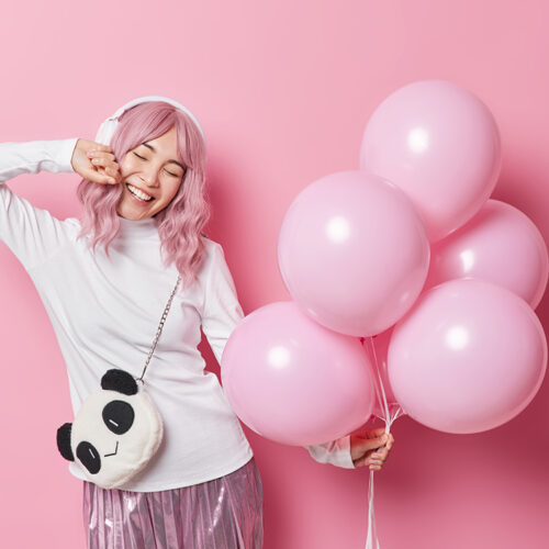 Positive long haired millennial girl listens favorite music holds bunch of inflated balloons carries bag wears white turtleneck and skirt has festive mood isolated on pink background. Holiday concept