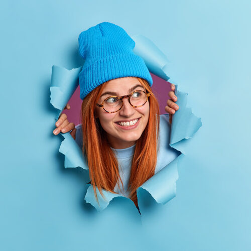 Positive redhead young woman looks away with pleasant smile has curious expression wears hat and optical glasses breaks through blue paper background. Millennial girl has foxy hair happy smile