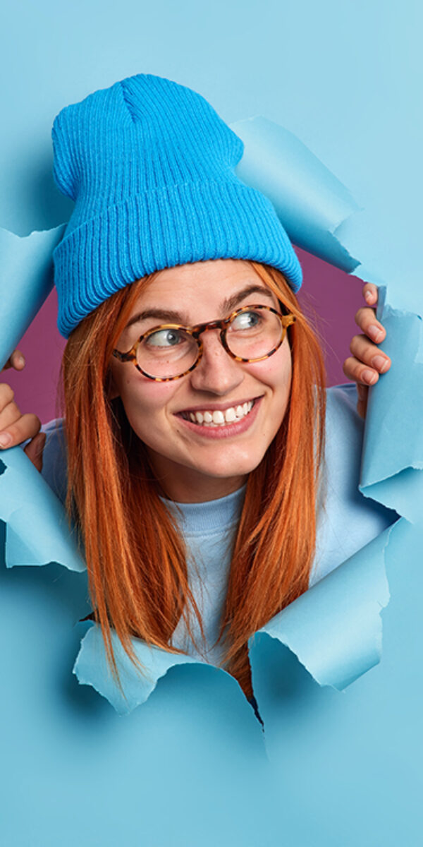 Positive redhead young woman looks away with pleasant smile has curious expression wears hat and optical glasses breaks through blue paper background. Millennial girl has foxy hair happy smile