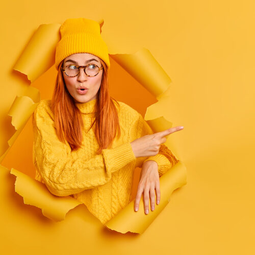 Pretty redhead gir with wondered expression indicates at blank space says wow looks at this promo wears hat and knitted sweater poses through hole of paper yellow background. Advertisement concept