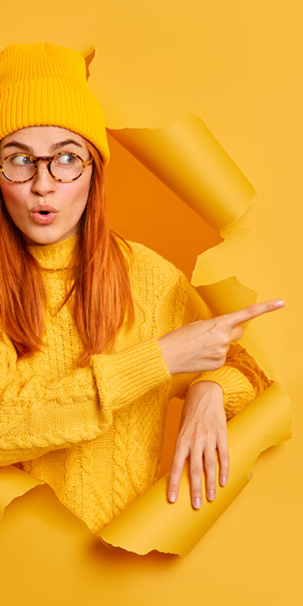 Pretty redhead gir with wondered expression indicates at blank space says wow looks at this promo wears hat and knitted sweater poses through hole of paper yellow background. Advertisement concept