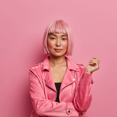 Serious stylish lady looks with calm expression direcrly at camera, has healthy skin, rouge cheeks, wears pink hair wig, dressed in jacket, keeps hand raised, isolated over rosy background. Monochrome