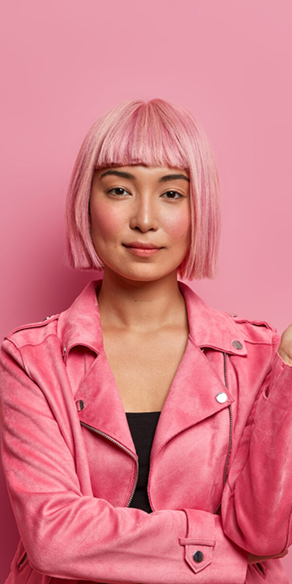 Serious stylish lady looks with calm expression direcrly at camera, has healthy skin, rouge cheeks, wears pink hair wig, dressed in jacket, keeps hand raised, isolated over rosy background. Monochrome