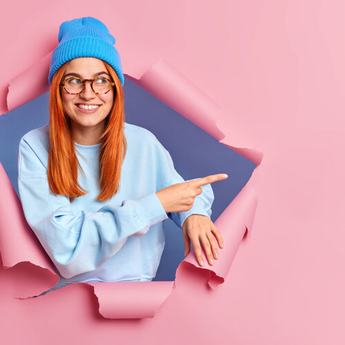 Smiling ginger woman feels optimistic points away on blank space shows advertisement wears blue hat glasses and jumper breaks through paper background promots some item. Place your promotion here