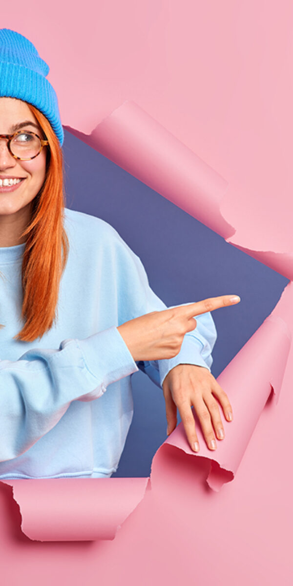 Smiling ginger woman feels optimistic points away on blank space shows advertisement wears blue hat glasses and jumper breaks through paper background promots some item. Place your promotion here