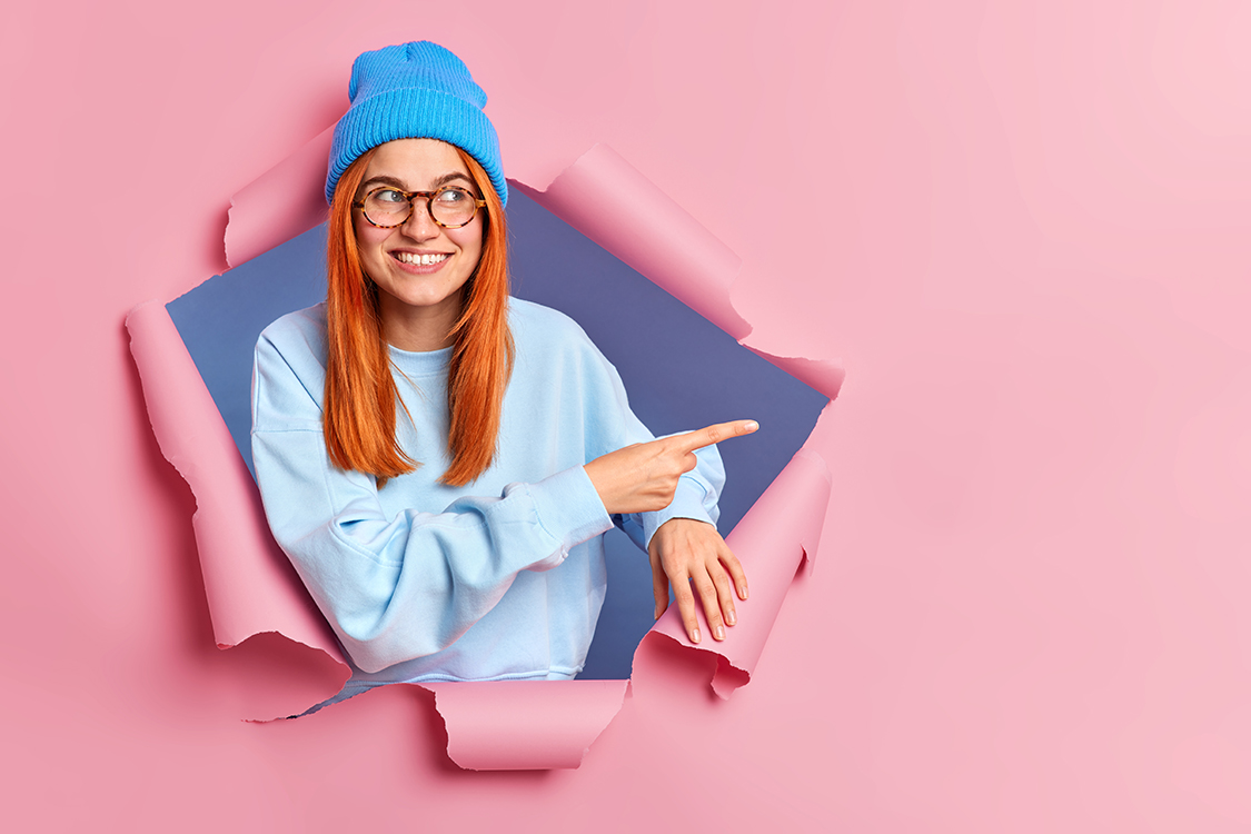 Smiling ginger woman feels optimistic points away on blank space shows advertisement wears blue hat glasses and jumper breaks through paper background promots some item. Place your promotion here