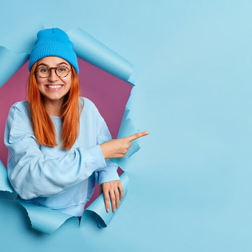 Smiling teenage girl with red hair gives recommendation or stands satisfied shows advertising content or big sale offer breaks through blue paper background. Empty space for your promotion here