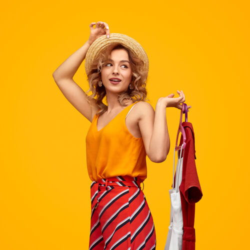 Smiling trendy blonde with trendy clothes Happy young woman in straw hat looking away and holding hangers with stylish garments against vivid yellow background