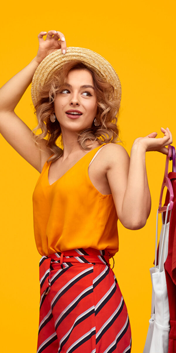 Happy young woman in straw hat looking away and holding hangers with stylish garments against vivid yellow background