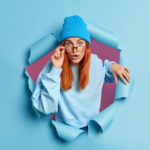 Surprised redhead European woman has shocked face expression stares with eyes popped out reacts on astounding news wears casual jumper and hat poses through ripped paper hole. Reaction concept