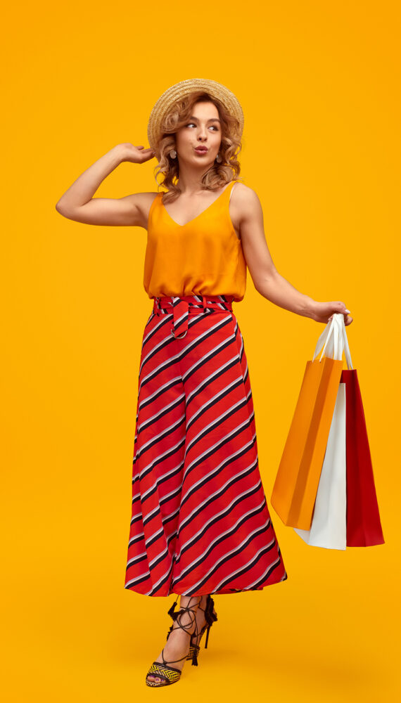 Full length stylish young female with paper bags pouting lips and looking away against yellow background