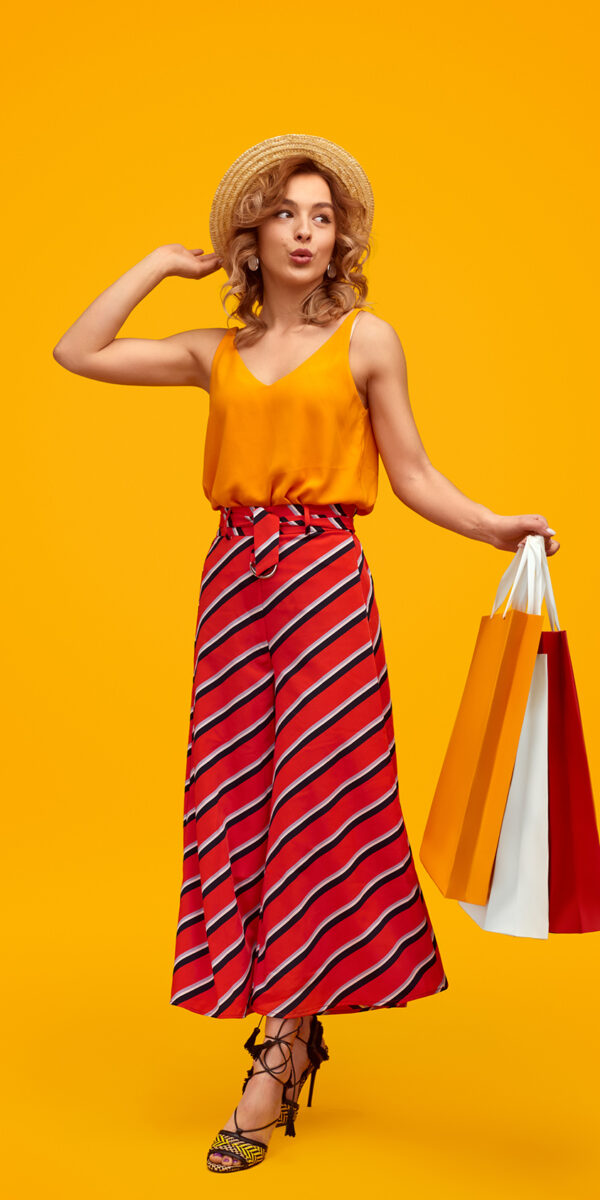 Full length stylish young female with paper bags pouting lips and looking away against yellow background