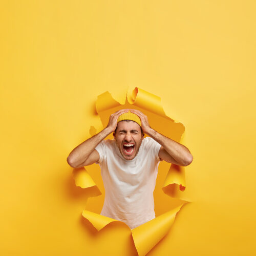 Vertical shot of emotional guy touches head with both hands, wears casual white t shirt, stylish yellow hat, poses in ripped hole paper, shouts from negative emotions, feels annoyed. Bad feelings