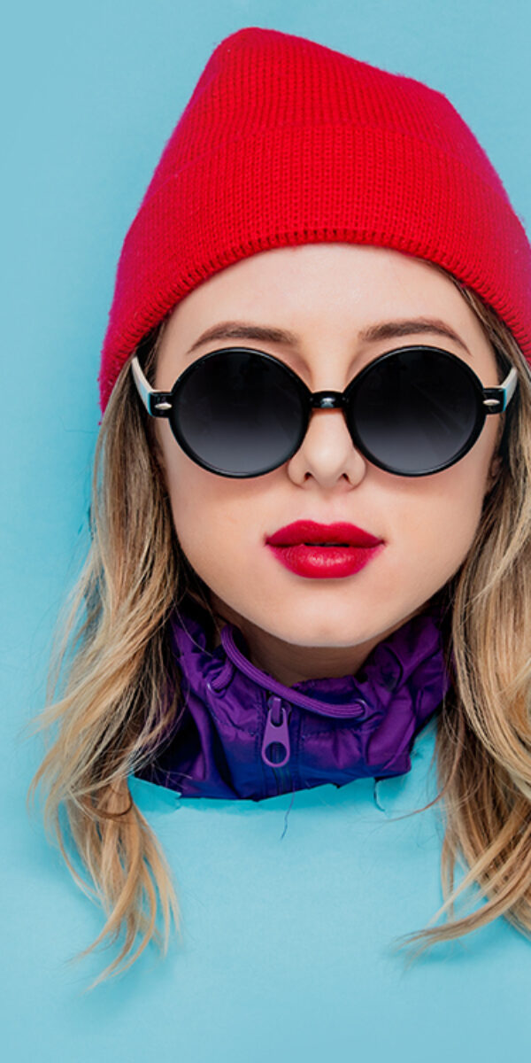 Portrait of a woman in red hat, sunglasses and suit of 90s on blue background.