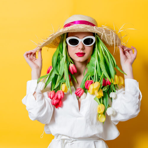 Beautiful woman in white shirt and hat with fresh springtime tulips and sunglasses on yellow background